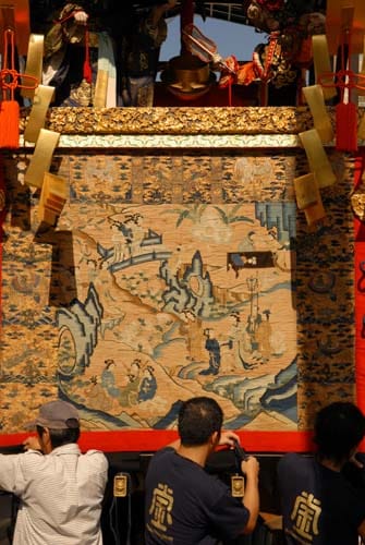 A vintage textile depicting Chinese court ladies decorates a Gion Matsuri float under the bright summer sun