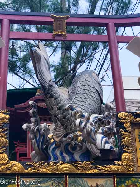 Wooden carving of a carp swimming up a waterfall through a red Shinto gate.